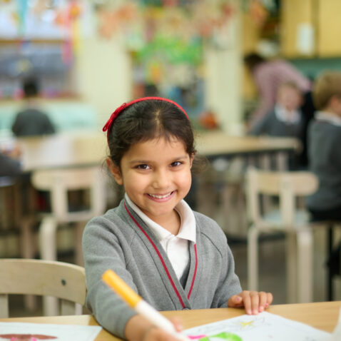 Smiling dark-haired schoolgirl, wearing a red hairband, is colouring in with a yellow felttip pen